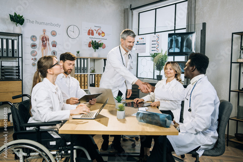 Bearded caucasian doctor shaking hands and greeting his multi ethnic colleagues during medical seminar at office. Female wheelchair user working with colleagues on common project.