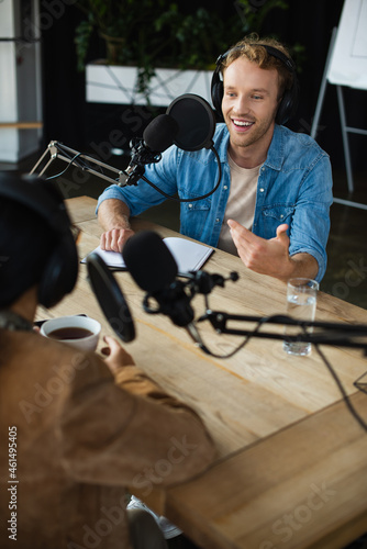 cheerful radio host in headphones looking at blurred colleague and gesturing during podcast