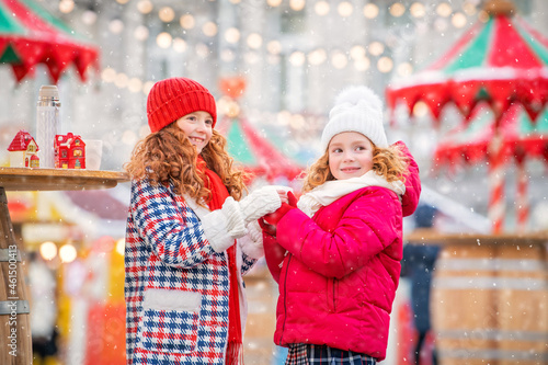 Children, red-haired sisters warm their gloved hands with a mug of hot tea at a festively decorated Christmas market in the city.