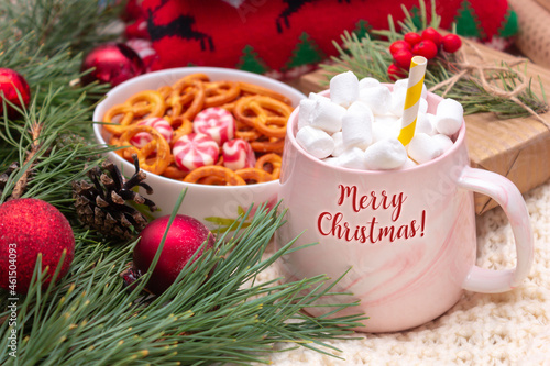 A mug with the text Merry Christmas with marshmallow and straw near a bowl of pretzels, a sweater, a gift box and branch of a Christmas tree on a white knitted plaid. New Year's holiday decorations