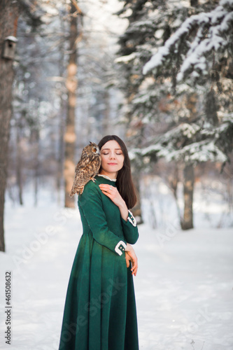 Friendship of an owl and a girl. Myth, fairy tale. Mystical spirit. Fantasy female elf walks with an owl in the winter forest. Beautiful brunette brunette girl in a green vintage dress. Tame bird.