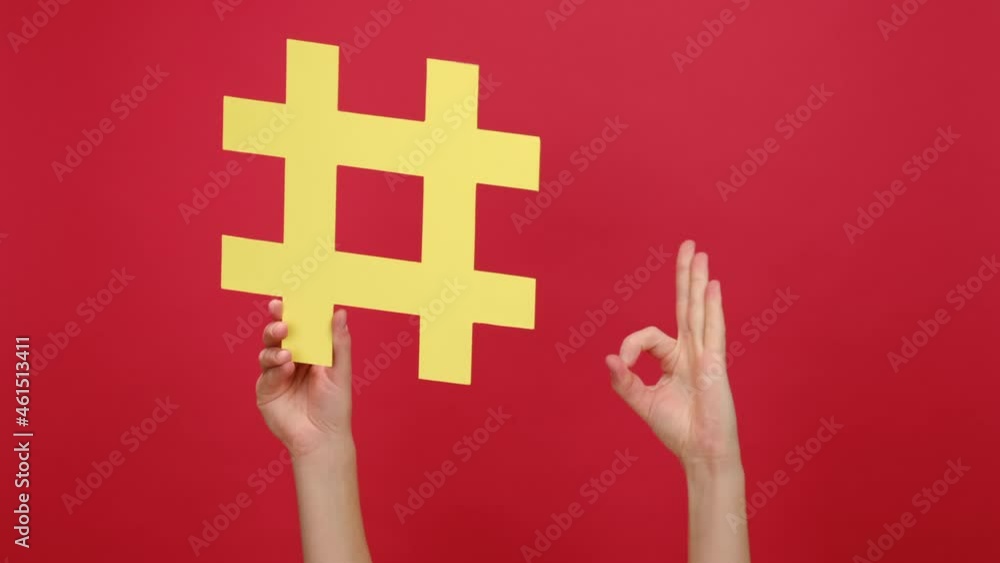 Female hand holding large yellow hashtag and pointing finger to it showing okay sign, isolated on red color background wall in studio. Social media marketing symbol, blogging, media planning concept