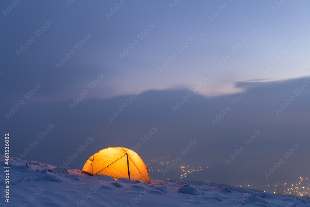 Yellow tent lighted from the inside against the backdrop of glowing city lights in fog. Amazing snowy landscape. Tourists camp in winter mountains. Travel concept