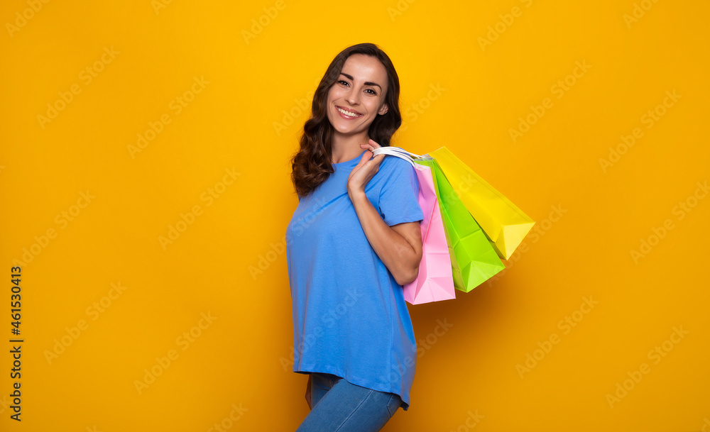 Happy smiling young woman in casual clothes with colorful shopping bags is posing on yellow background Happy smiling young woman in casual clothes with colorful shopping bags is posing on yellow background