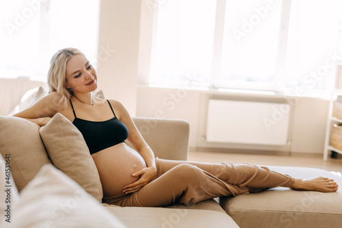 Pregnant woman wearing cozy home wear, lying on a sofa in a light interior.