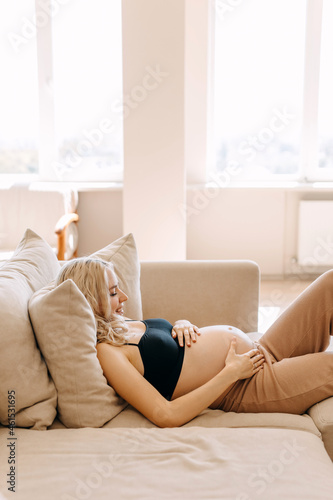 Pregnant woman wearing cozy home wear, lying on a sofa in a light interior, holding hands on belly.
