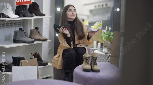 Young millennial woman choosing between yellow casual sneakers and stylish elegant green high-heels sitting in shoe store. Wide shot portrait of thoughtful Caucasian buyer making decision. Style photo