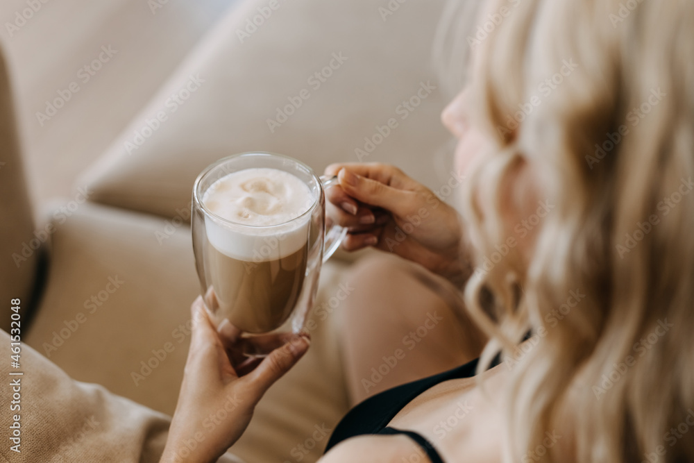 Closeup of a woman holding a double glass cup of coffee, sitting on sofa at home.