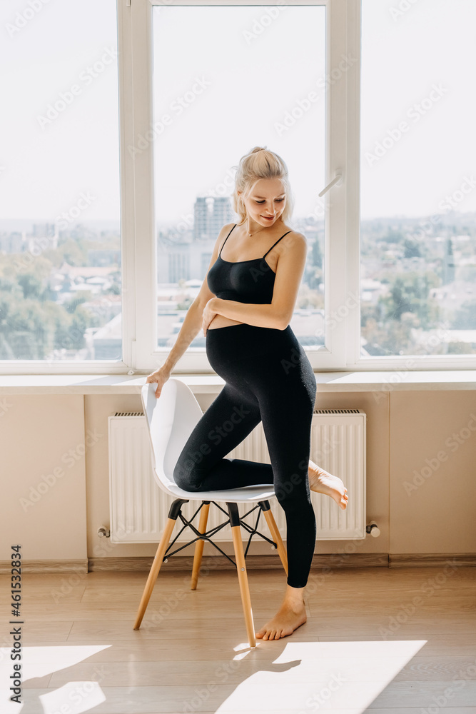 Blonde pregnant woman wearing black top and leggins, standing next to a chair at home. Blonde pregnant woman wearing black top and leggins, standing next to a chair at home.