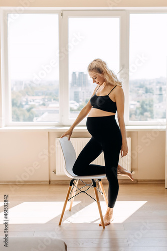 Blonde pregnant woman wearing black top and leggins, standing next to a chair at home, looking at her belly.