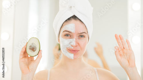 A woman with a towel on her hair and in a clay face mask holds an avocado. Taking care of beauty at home photo