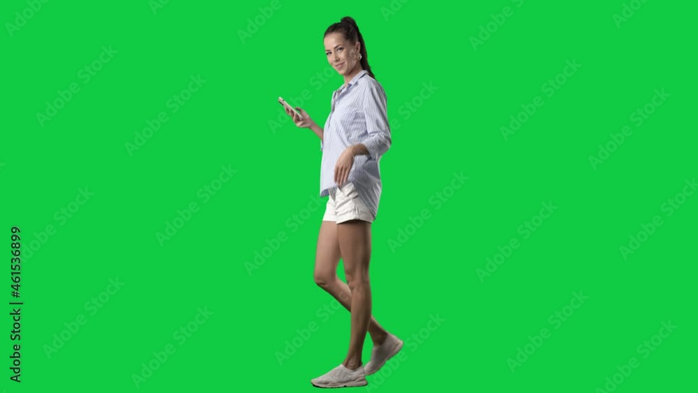 Side view of stylish summer girl walking using phone turning at camera greet and wave hand. Full body on green screen chroma key background Side view of stylish summer girl walking using phone turning at camera greet and wave hand. Full body on green screen chroma key background