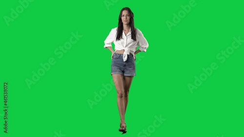 Confident attractive brunette woman walking wearing white tied shirt and jeans short. Front view. Full body on green screen chroma key background