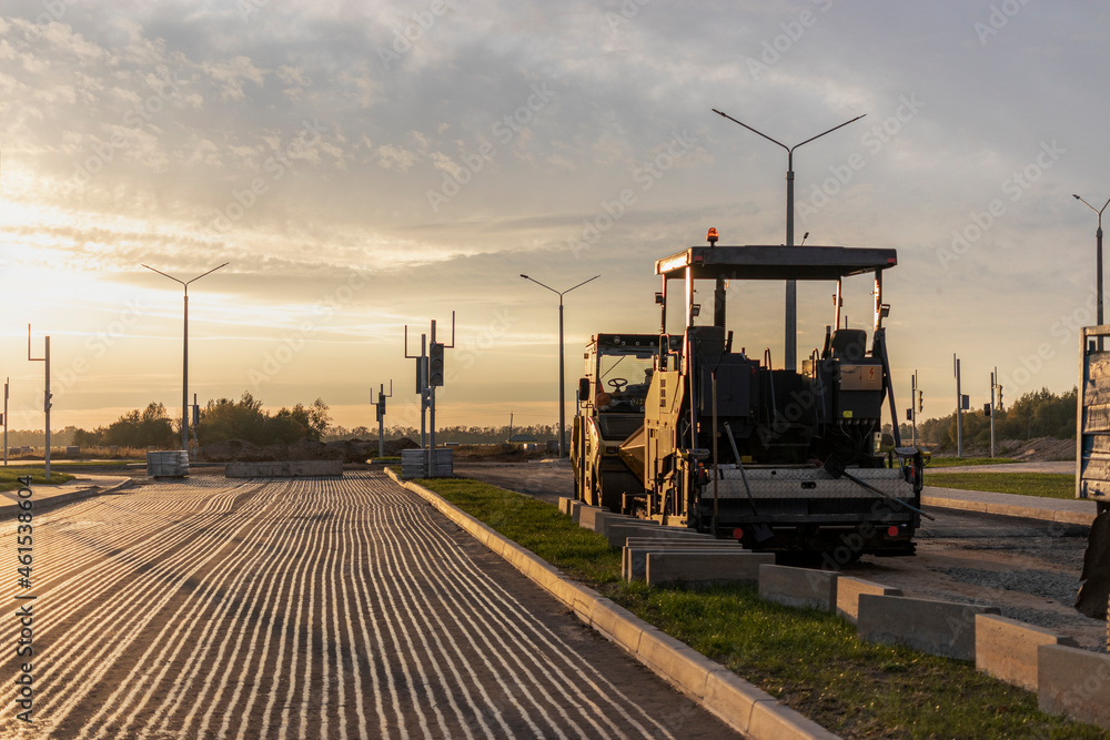 Road repair and asphalt paving with a specialized machine in the evening against the backdrop of the sunset sky. The afalt paver works in the evening repairing the road surface. Road repair and asphalt paving with a specialized machine in the evening against the backdrop of the sunset sky. The afalt paver works in the evening repairing the road surface.