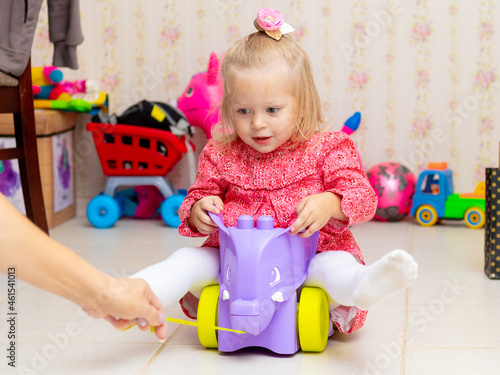 A little blond girl rides a toy car while playing