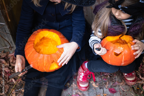 Little girls make jack-o-lantern from big pumpkins for celebratiion of halloween holiday.Witch costume, hat, coat. Cut with knife,take out pulp with seeds.Outdoors activity, backyard.Children's party photo