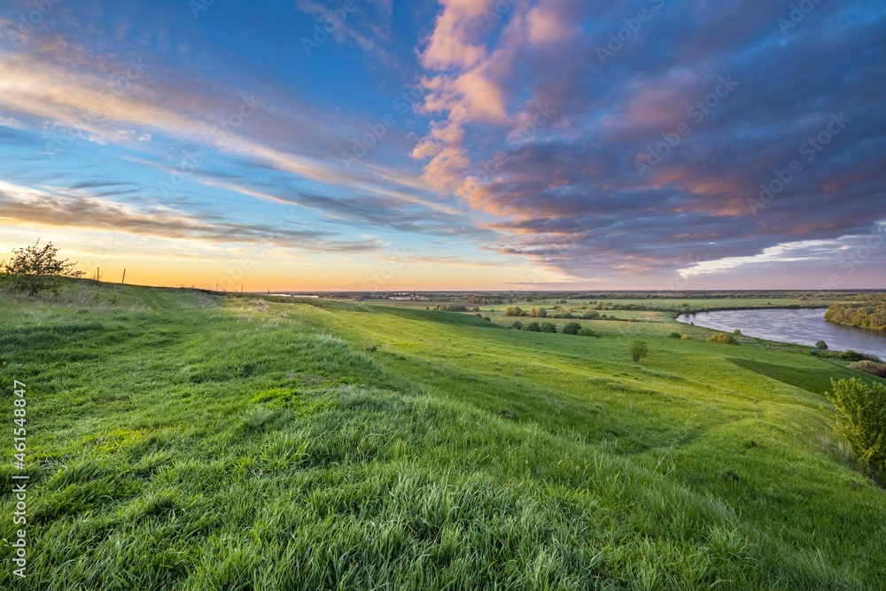 Obraz premium Scenic view of a beautiful summer river sunset with reflection in the water, calm water. Bright colorful cloudy sky. Lush green grass in the foreground.