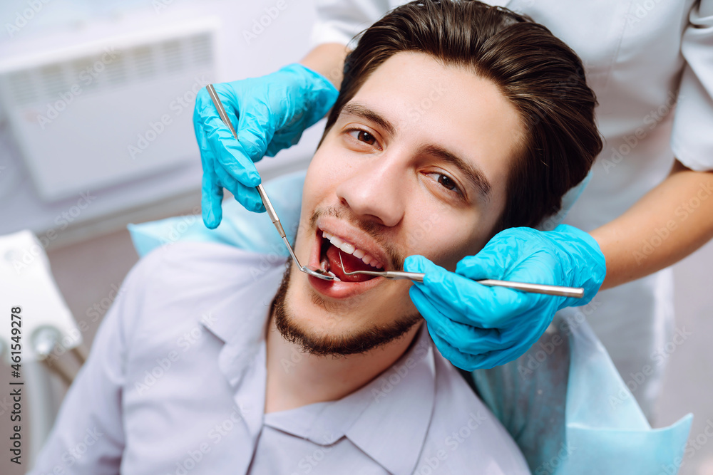 Young man at the dentist's chair during a dental procedure. Overview of dental caries prevention. Dentist examining patient's teeth in modern clinic. Healthy teeth and medicine concept. Young man at the dentist's chair during a dental procedure. Overview of dental caries prevention. Dentist examining patient's teeth in modern clinic. Healthy teeth and medicine concept.