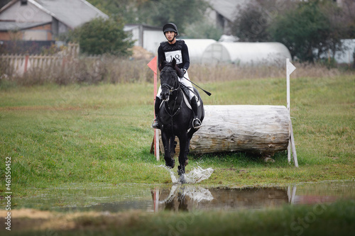 rider man galloping fast in water pond on black stallion horse during eventing cross country competition in autumn