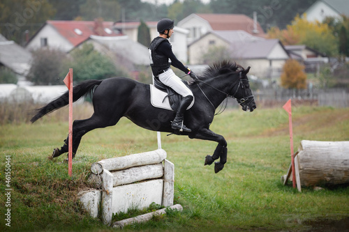 close portrait of handsome rider man jumping drop fence obstacle on black stallion horse during eventing cross country competition in autumn