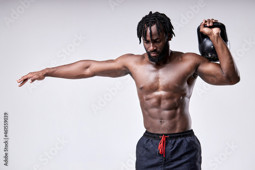 Black young fit muscular man training with kettlebell isolated on white background. Strength and motivation, sport concept. Strong shirtless guy raising kettlebell, having good physical shape.
