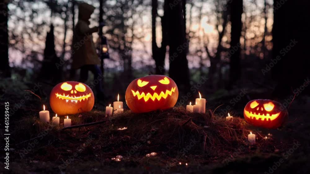 Mysterious and creepy Halloween concept. Glowing jack-o-lantern pumpkins and burning candles at night. Unrecognizable brave little girl with lantern walking through dark forest in background Mysterious and creepy Halloween concept. Glowing jack-o-lantern pumpkins and burning candles at night. Unrecognizable brave little girl with lantern walking through dark forest in background