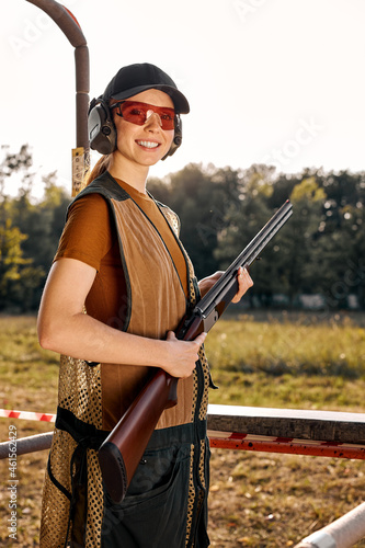 Smiling caucasian woman trains to shoot with double-barreled hunting rifle on sports stand, attractive lady posing with rifle, wearing uniform, protective eyeglasses and cap. outdoor portrait