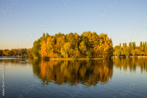 A small island with trees with thick multicolored foliage on a pond with a reflection on a clear sunny October day in Izmailovsky Park in Moscow and a space for copying