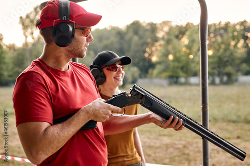 Handsome male shooting instructor teaches the backslider how to handle weapon on training course in outdoor range. positive teacher and woman in protective spectacles and headset have fun, laugh