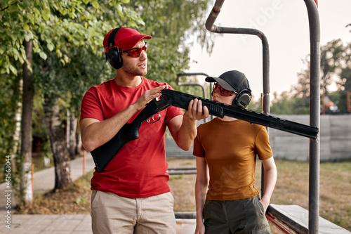 handsome confident male teach female to recharge a rifle on shooting range outdoors, give instructions and have talk, wearing protective headset, spectacles and cap, holding rifle in hands.
