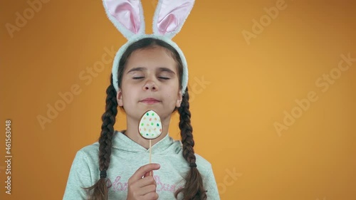 Happy Easter. Beautiful baby girl eats egg on a stick on a yellow background. Kid with rabbit ears eating Easter cookies.