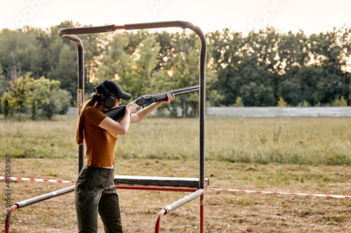 Fototapeta young caucasian woman in goggles and headset aiming rifle at side, ready to shoot in an outdoor range