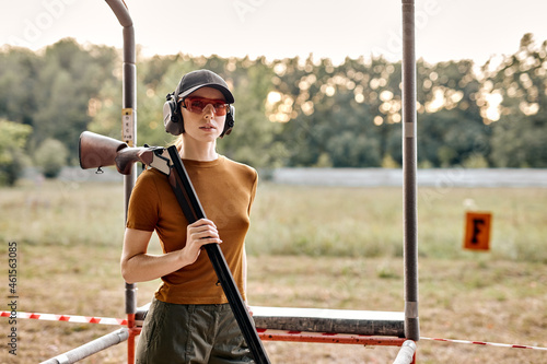 Portrait of young caucasian lady in cap after shooting practice on outdoor range, holding rifle, training alone, dressed in casual t-shirt, cap, protective eyeglasses and headset, look at camera