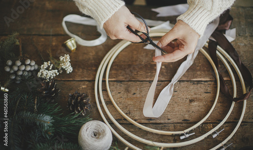 Making stylish minimalist christmas wreath. Hands cutting ribbon with scissors for modern boho wreath with fir branches, brunia herb, wooden hoop on rustic table. Atmospheric moody image