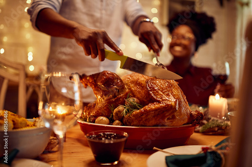 Close-up of African American man carves roasted Thanksgiving turkey at dining table.