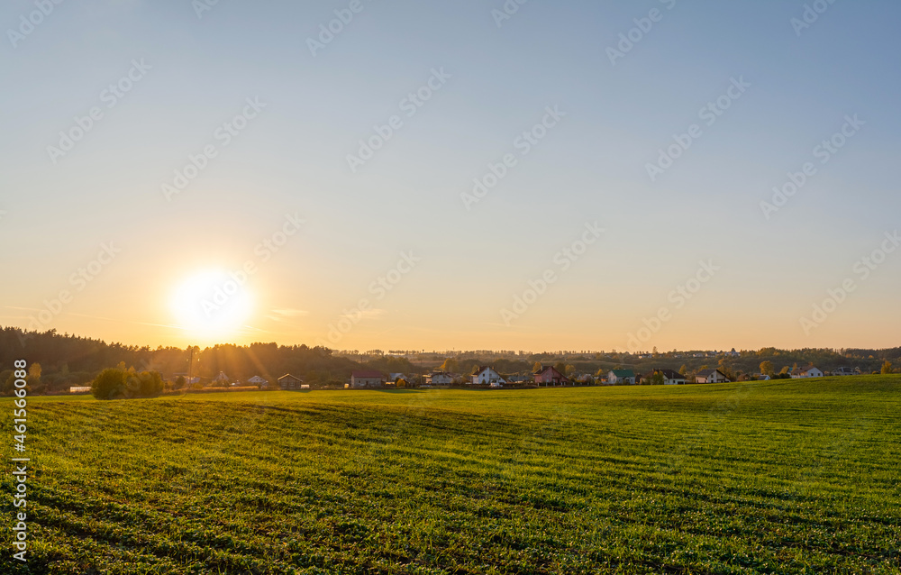 Green agricultural field, holiday village on the horizon. Blue orange sky during sunset. Autumn Nature