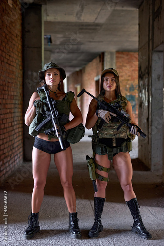 Two Military Female Soldiers in Gear Ammunition Posing At Camera With Weapons Guns. Sexual Members Of Military Forces Ready To Strike, Country Defense On Battle Fight Area, In Building