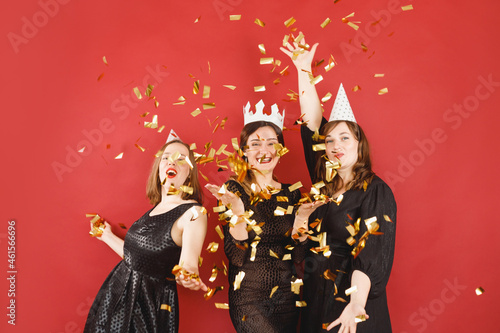 Three delighted women in black dress and party hats over red background with golden confetti. Female party