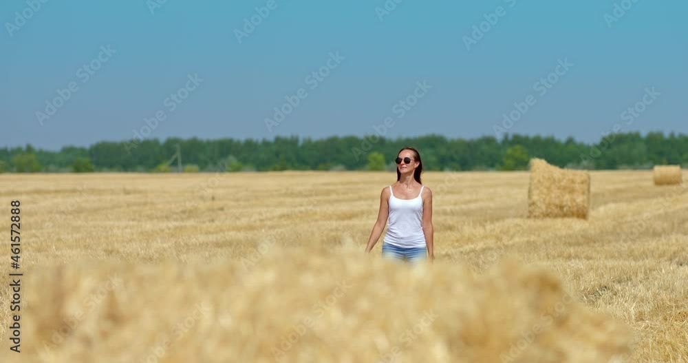 Happy woman in jeans and sunglasses walks through a field near a large golden haystack. Walk in a wheat field with mown grass, haystacks lying in the sun. Beautiful rural landscape. 4k, ProRes Happy woman in jeans and sunglasses walks through a field near a large golden haystack. Walk in a wheat field with mown grass, haystacks lying in the sun. Beautiful rural landscape. 4k, ProRes