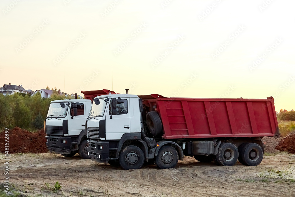 Two red dump trucks simultaneously lifted the bodies to unload the sand. Cargo transportation services. Large multi-ton truck. Unloading cargo. Construction site and machinery. Banner. Common view