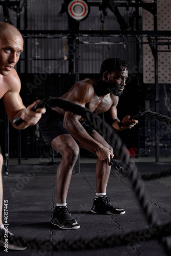 two diverse athletic guys in dark cross fit class, males started training exercises get sweating, holding battle ropes in hands, concentrated and motivated. sportive active healthy lifestyle.