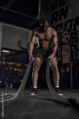 Athletic active african man with battle rope doing exercise in functional training fitness gym exercising with battle ropes at gym. young shirtless guy working out arms and cardio for cross fit