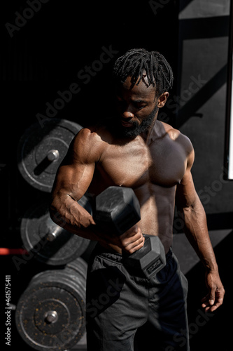 active strong sweaty focused fit muscular man with big muscles holding heavy dumbbell for cross fit training, black man do hard core workout in dark modern gym, real people exercising. portrait