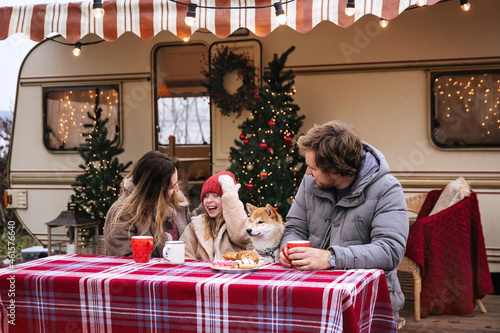 Celebrating Christmas in camper: young family drinking hot cocoa sitting outside Rv van decorated for Xmas celebration. Parents and daughter on road trip on winter holidays or vacation outside city
