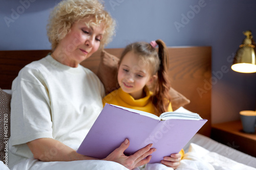 nice grandmother reads a book to little daughter lying on bed in cozy children's room, domestic atmosphere. friendly aged woman enjoys spending time with grandchildren, relaxing together