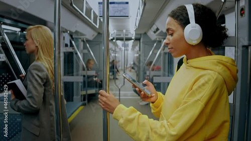 A young girl standing in modern metro train, wearing headphones and looking in her phone. A man walks past her, obscuring the frame. She is wearing a bright yellow hoodie and white headphones.  photo