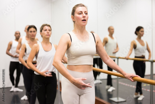Group of people doing ballet exercises using barre in gym with focus to fit athletic toned woman in foreground in health and fitness concept.