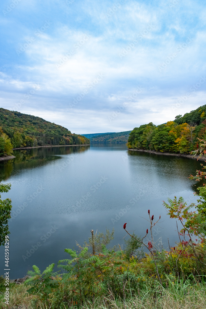 Fototapeta premium Pepacton Reservoir, Catskills, NY. Beautiful calm lake surrounded by forest. Colors of the leaves starting to change with the fall season. Selective focus on background. Blue sky, copy space.