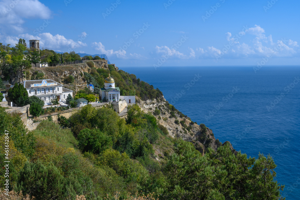 View of the Black Sea and the ancient monastery on a picturesque rock covered with green plants on a sunny summer day. Golden domes glisten in the sun. Blue sky with white clouds. Crimea 