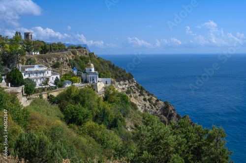 View of the Black Sea and the ancient monastery on a picturesque rock covered with green plants on a sunny summer day. Golden domes glisten in the sun. Blue sky with white clouds. Crimea 